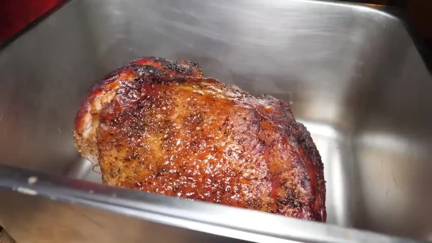 High-angle shot of a chef wearing black professional gloves shredding a large, tender piece of slow-smoked barbecue pork shoulder in a silver pan. The footage highlights the succulent, juicy texture o
