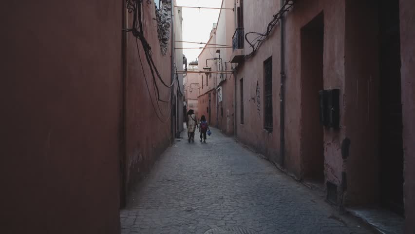 A frontal view of a narrow Moroccan alley with warm terracotta walls, two people walking away toward soft daylight filtering through the passage