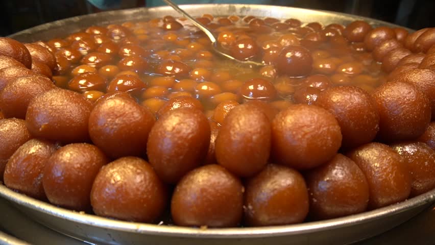 A large metal tray filled with sweet brown gulab jamun balls completely soaked in sugary syrup at a sweet shop