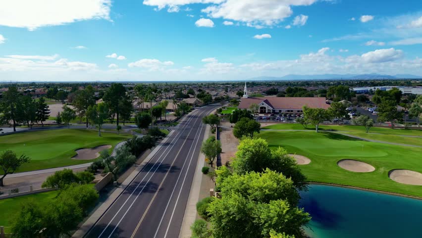 An aerial view of a sunny Mesa, Arizona roadway bordered by lush golf greens, tall trees, and a nearby community church under bright blue sky
