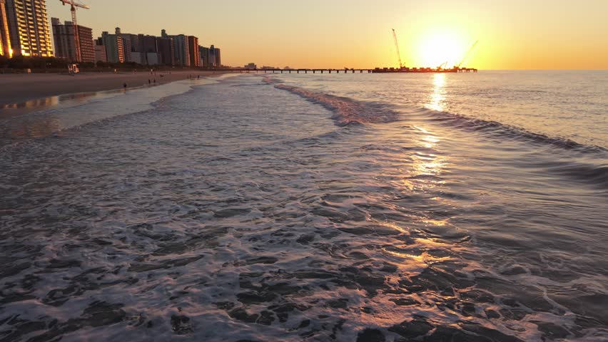 Waves gently lap against the shore at sunset, with beachfront buildings and a pier visible in the background at Myrtle Beach, South Carolina