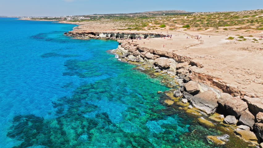 Aerial view of Cape Cavo Greco sea caves in Cyprus. Tourists visit the rocky cliff area above crystal-clear turquoise water, natural cave openings and rugged Mediterranean coastline.