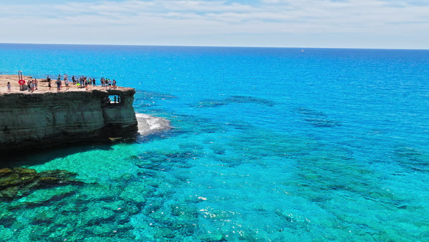 Aerial view of Cape Cavo Greco sea caves in Cyprus. Tourists visit the rocky cliff edge above crystal-clear turquoise water, with natural cave openings, limestone cliffs and the open Mediterranean Sea.