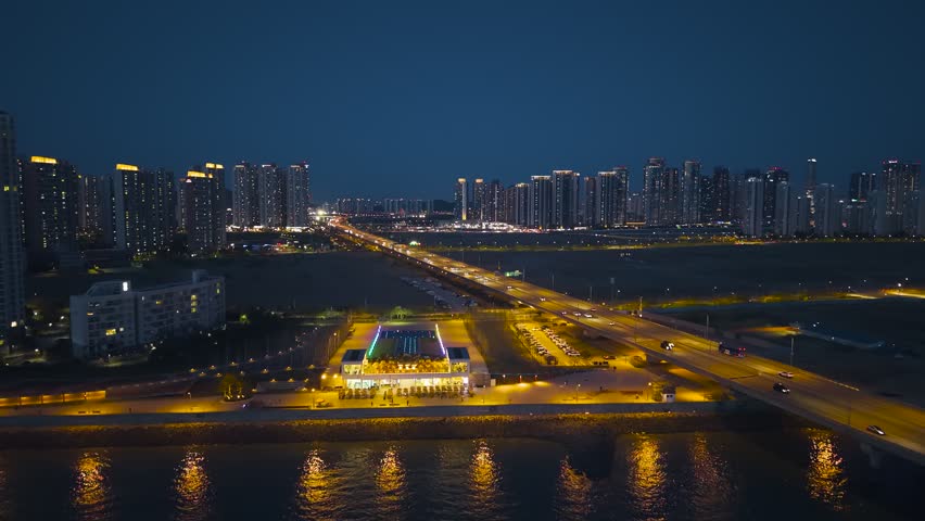 Aerial flypast reveals illuminated seafront cafe rooftop deck and night traffic crossing Incheon Bridge expressway toward Songdo skyline