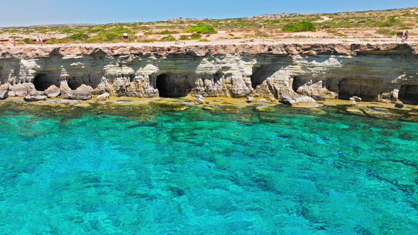 Aerial view of Cape Cavo Greco sea caves in Cyprus. Tourists visit the rocky shore beside turquoise water, cave openings and dramatic Mediterranean cliffs.