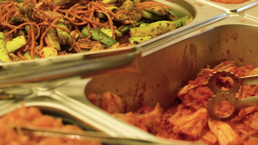 Close-up shot of metal tongs serving fermented vegetables from a stainless steel buffet tray.