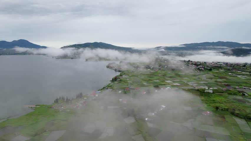 Aerial drone footage of low clouds drifting over a tranquil mountain lake, patchwork farmland, and a rural village surrounded by lush green hills during an early morning landscape.