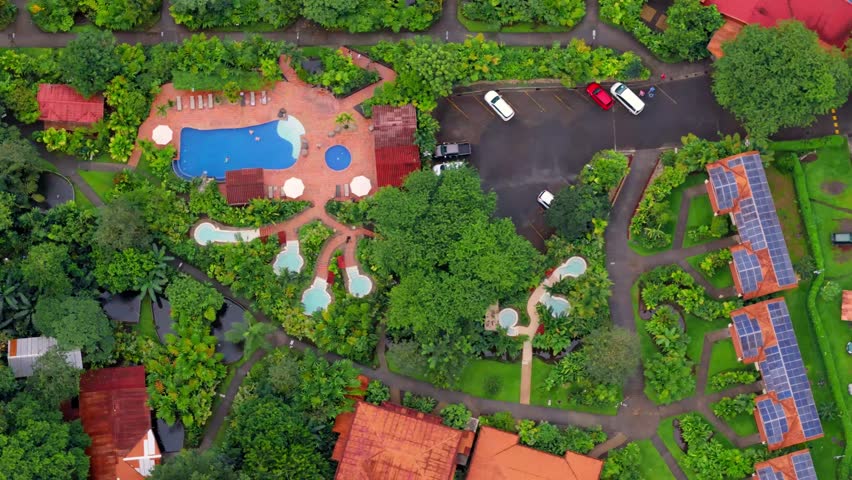 Soaring aerial perspective revealing a beautiful eco-friendly resort nestled in lush green vegetation, featuring swimming pools, hot tubs, and bungalows equipped with modern solar panels