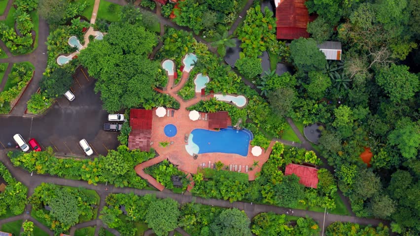 Aerial top-down view of a luxury eco-resort nestled in a lush green jungle, featuring beautiful swimming pools, bungalows with solar panels, and serene walking paths