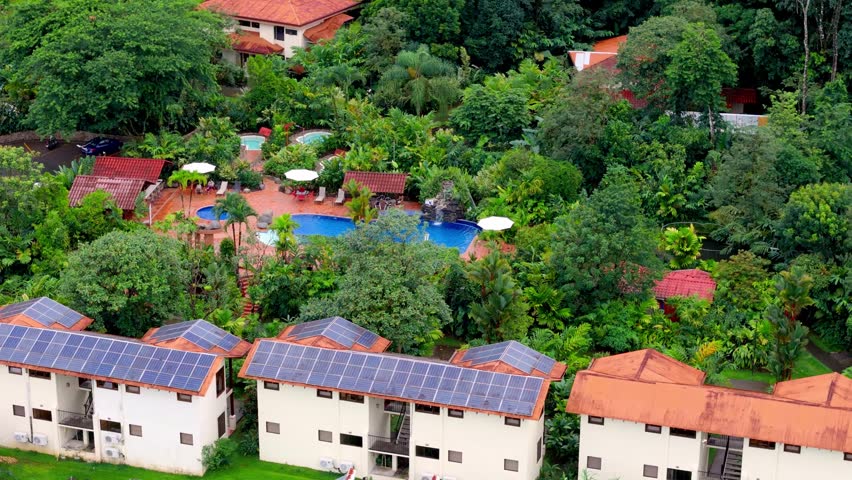 High-angle view showcasing a sustainable eco-friendly resort with solar panels installed on the roofs of its buildings, surrounded by dense green tropical vegetation and a swimming pool area