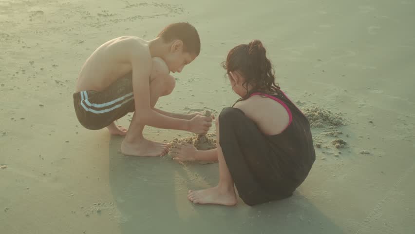 Two children sit together on the sandy beach, focused on playing and creating shapes in the sand. A warm family travel moment that captures curiosity, creativity, and childhood exploration.