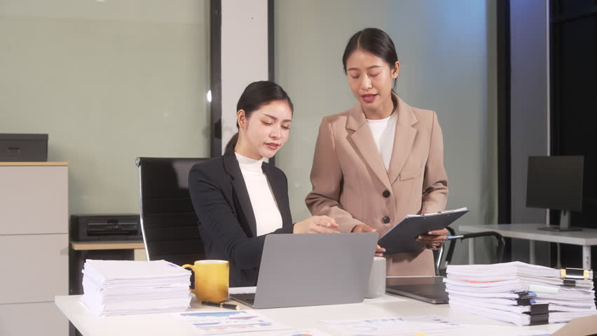 Two businesswomen reviewing documents and working on a laptop in a modern office