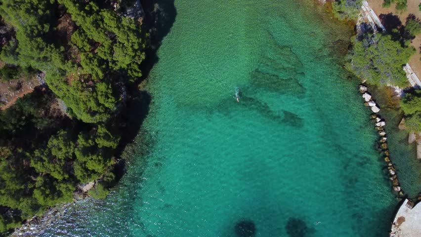 Top down aerial view of a person swimming in pristine turquoise water. People are sunbathing and relaxing on the small pebble beach of a secluded cove