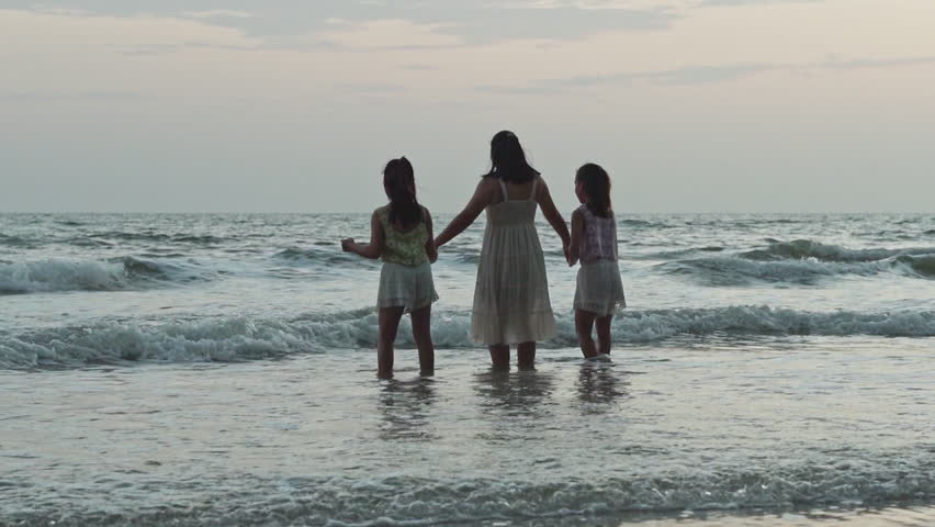 Happy mother and two daughters holding hands, running, jumping and playing together in ocean waves on beach during summer vacation