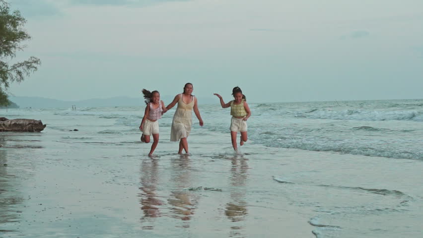 Happy mother and two daughters holding hands, running, jumping and playing together in ocean waves on beach during summer vacation