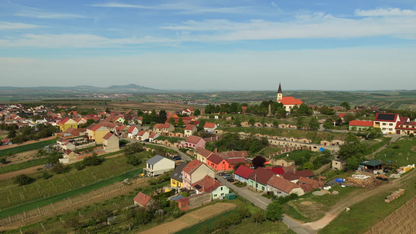 Aerial drone view of a small Czech village with a church steeple and colorful red-roofed houses surrounded by vineyards and rolling agricultural plains, Moravia, Czech Republic.