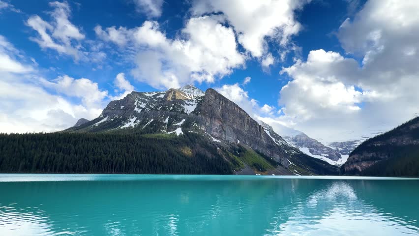 Time Lapse. A stunning view of the mountains surrounding Lake Louise in Alberta. The water reflects the sky and nearby trees in a bright blue color. Clouds float above the peaks.