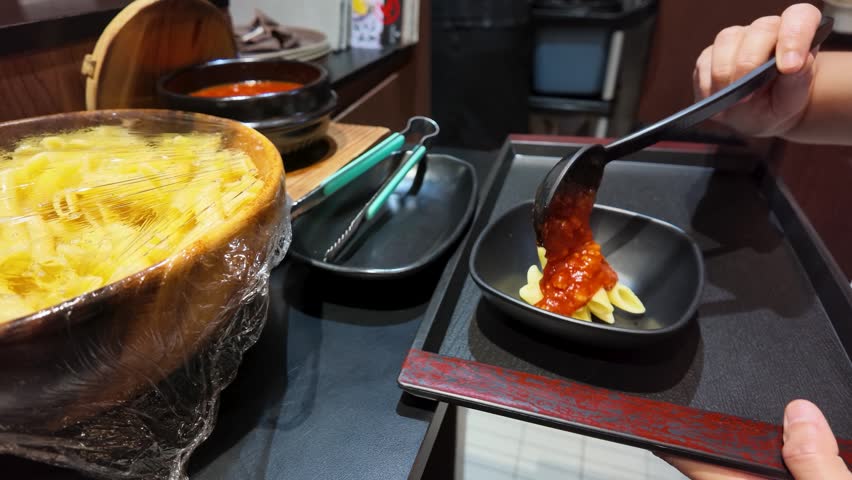 A person ladles red sauce onto penne pasta in a small bowl at a restaurant.