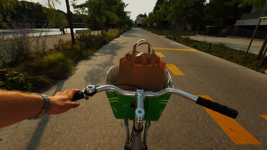 POV view of a person riding a city bike beside a river, with a reusable shopping bag in the front basket and warm evening light along the path.
