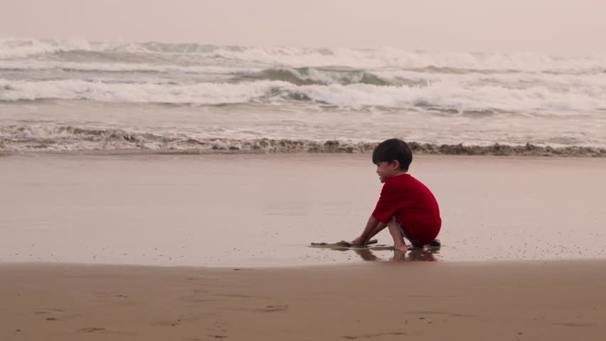 Child in red shirt sits barefoot while playing on the sand, Sunset bathes beach in soft, warm tones
