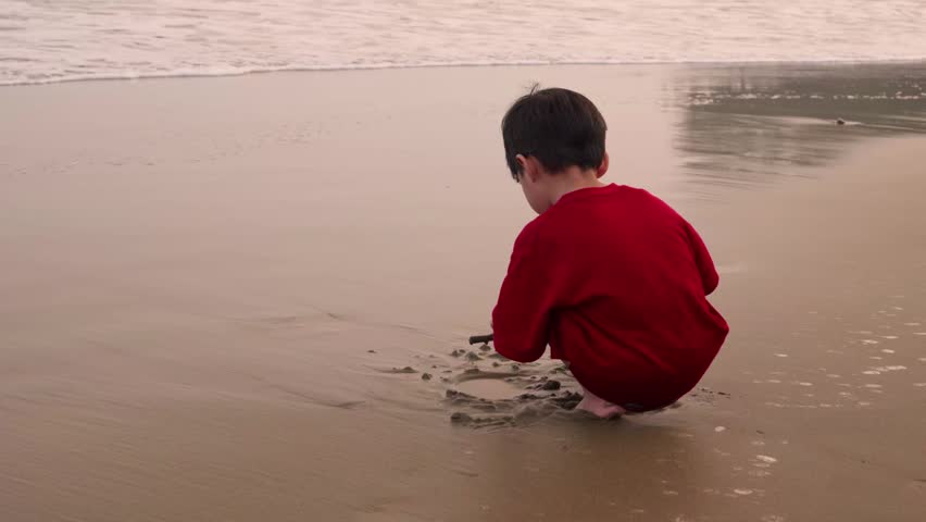Child in red shirt sits barefoot while playing on the sand, Sunset bathes beach in soft, warm tones