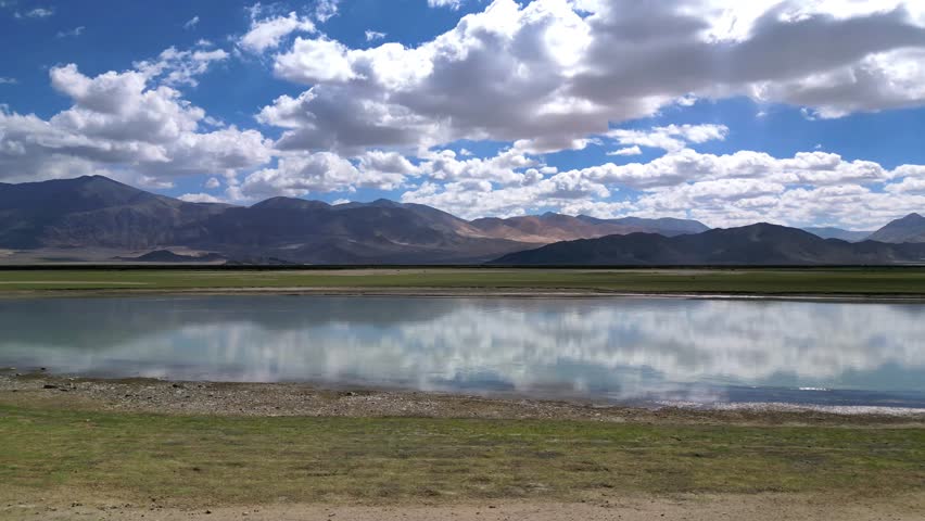 Aerial view of the lake in Dungti, surrounded by grassy banks and the rugged mountains of Ladakh under a blue sky with scattered clouds in Dungti, Ladakh, India.