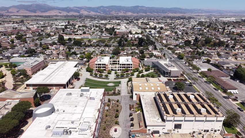 Aerial view of Hartnell College in Salinas, California, showcasing modern campus buildings, athletic facilities, blue pool, tree-lined walkways, and city backdrop under bright daylight. Pull out shot