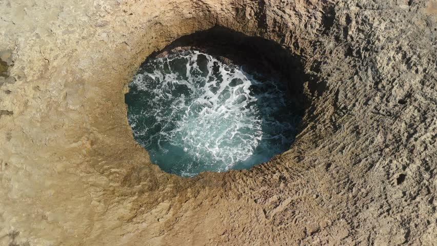 Aerial parallax circles Watamula Hole in Curacao showing waves surging through natural rock blowhole with faint rainbow mist