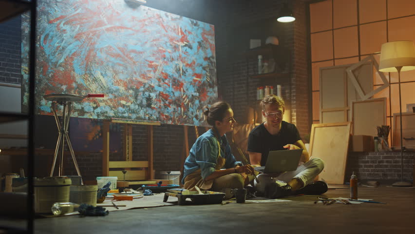 Couple of Artists Sitting in their Studio Use Laptop Computer, Working on a Project, Talking and Smiling. Painter and Her Partner Doing Research. Authentic Workshop with Oil Paintings and Tools
