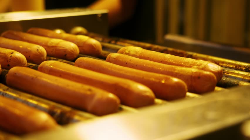 Hot dog sausages rotate on grill during cooking process. Street food vendor prepares grilled sausages for fresh takeaway orders.