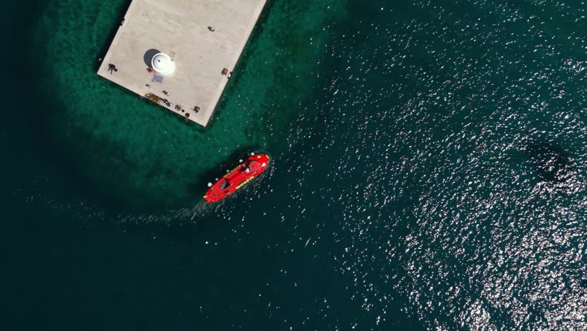 Red semi-submarine boat docking at a concrete pier with a lighthouse in Zadar, Croatia, top-down view.