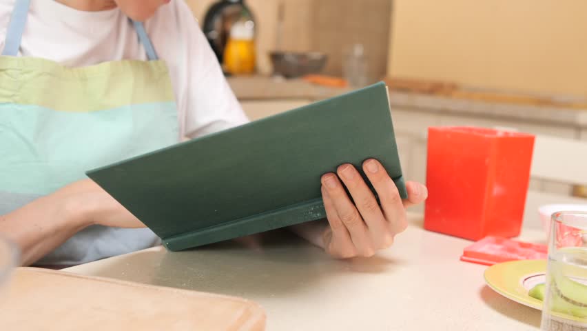 Person wearing an apron and holding a cookbook, focused on reading a recipe in a home kitchen, preparing for a cooking session