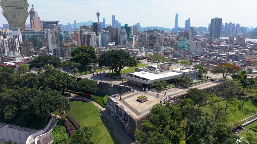 Aerial orbit reveals Macau Museum and Mount Fortress walls with tourists walking as Macau Tower and dense skyline rise in background