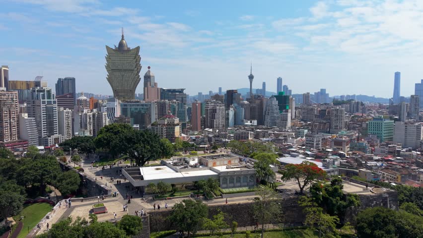 Golden Grand Lisboa tower and distant Macau Tower rise above Macau Museum and stone walls of Mount Fortress with tourists below - aerial parallax shot