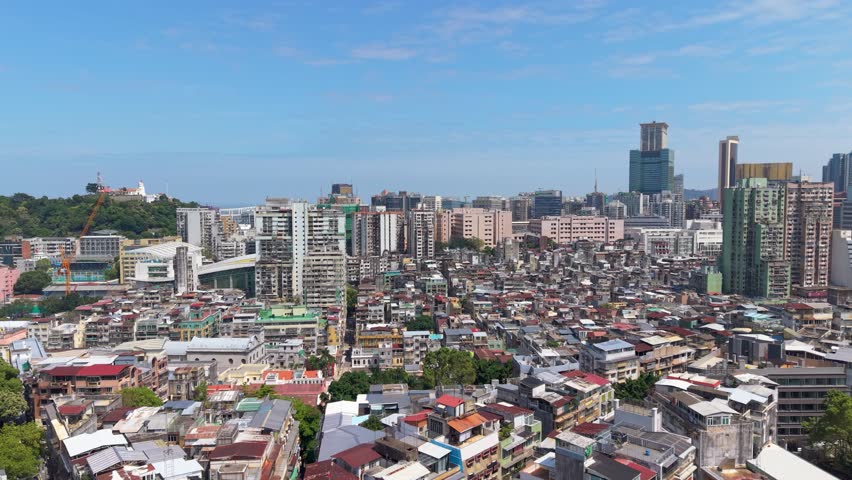 Forward flight over dense old town rooftops in Macau reveals skyline with Guia Fortress and Lighthouse on green hill beside modern skyscrapers
