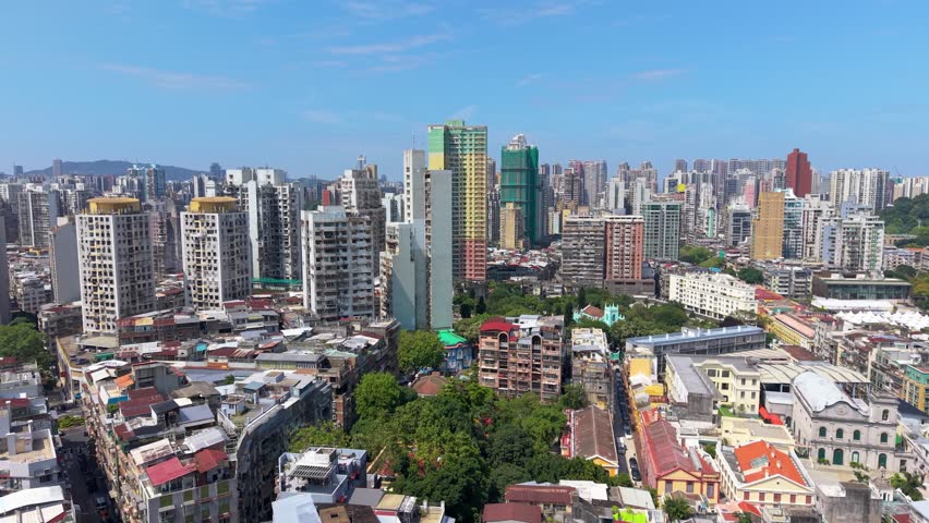 Parallax flight over dense Macau residential towers reveals crowded rooftops, green treetops, turquoise church spire and hazy hills beyond