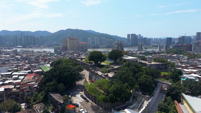 Orbiting aerial view of historic Mount Fortress walls and Macau Museum with tourists walking above river separating Macau from Zhuhai skyline