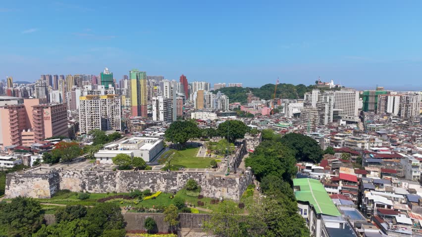 Parallax aerial reveals stone walls of Mount Fortress and Macau Museum courtyard with tourists strolling green lawn against dense city skyline