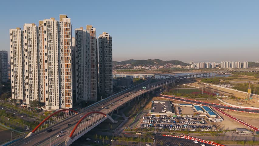 Aerial view captures Songdo apartment towers beside red arched bridge with cars crossing expressway interchange over river and parking lots