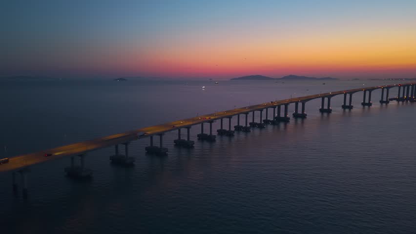 Aerial pan reveals Incheon Bridge cable towers silhouetted against pink orange blue hour sky with calm sea and distant island hills