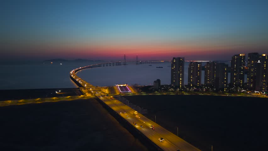 Aerial blue hour reveal of illuminated Incheon Bridge curving over water with expressway traffic lights beside glowing Songdo apartment towers