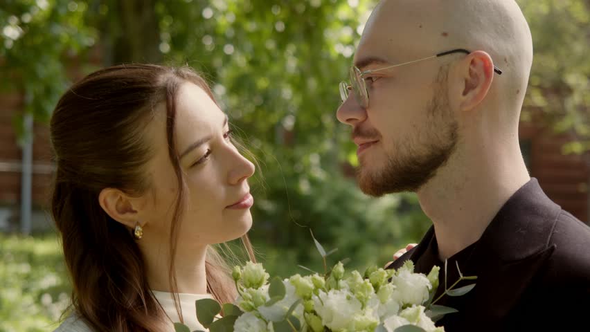 Newlywed couple kissing outdoors in warm summer light while holding wedding bouquet. Romantic close up wedding moment in natural garden setting.