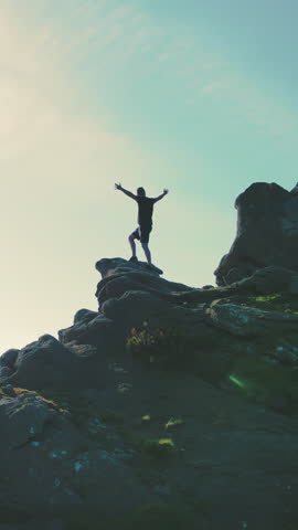 A man standing on top of a mountain with and raising his hands up the mountain. Victory and overcoming the top. A tourist looking at the sunrise on top of a rock.