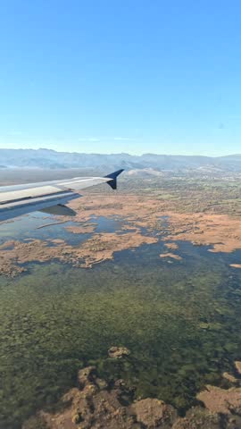 Airplane Window View Over Lake and Mountains. View from airplane window over lake, wetlands and mountains during flight. Aircraft wing, blue sky and aerial landscape from passenger seat.
