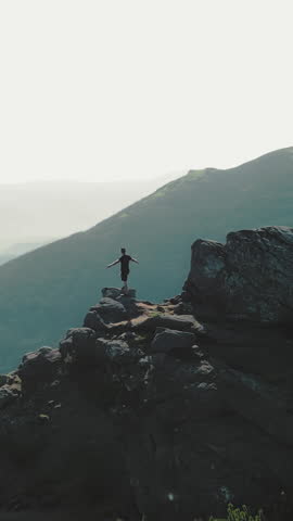 A man standing on top of a mountain with and raising his hands up the mountain. Victory and overcoming the top. A tourist looking at the sunrise on top of a rock.