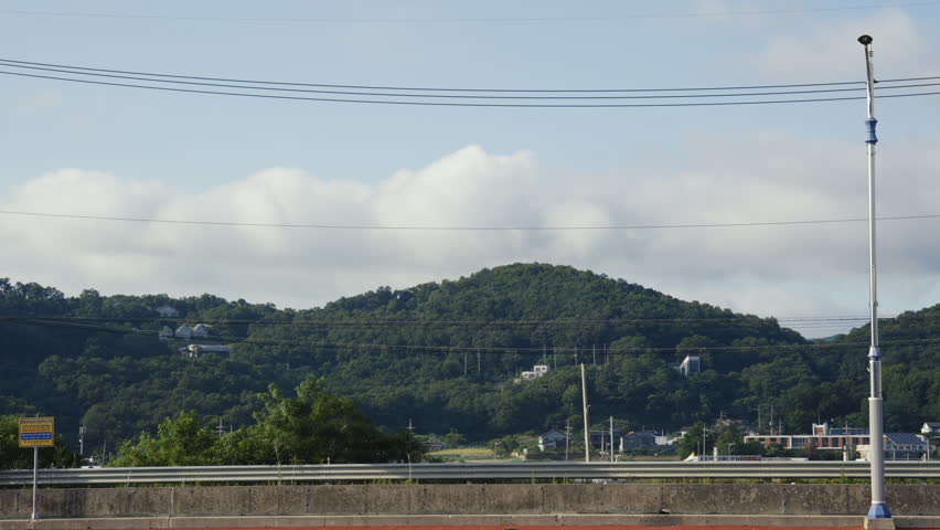 Numerous cars drive on an urban bridge; lush green hills stand against a blue sky