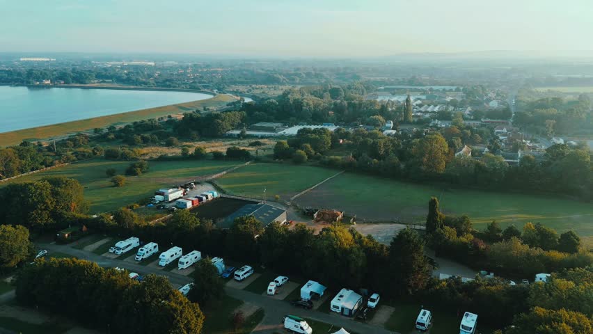 A drone aerial view of caravans and campers near King George V Reservoir, showing open fields, water edges, and morning haze across Chigwell, Essex