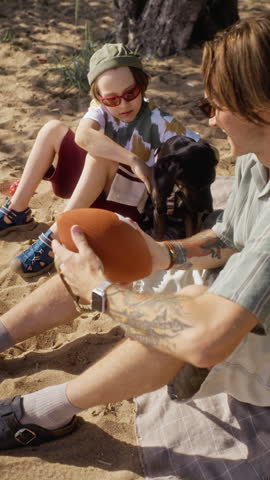 Vertical shot of Caucasian father and young child sitting on beach blanket with dog, playing with football. Ideal for family lifestyle marketing, pet care, or outdoor recreation campaigns