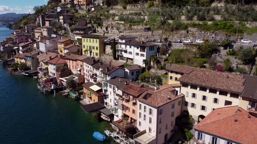 Colorful lakeside houses cling to steep terraced hillside in Gandria beside Lake Lugano, Switzerland, small wooden boats dock along waterfront near green olive terraces. Aerial drone orbit shot