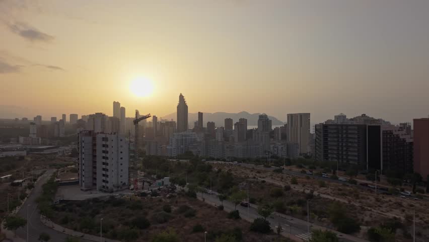 The sun sets over the city skyline as buildings stand tall. The sky shows orange and yellow colors, marking the end of the day during late afternoon hours.